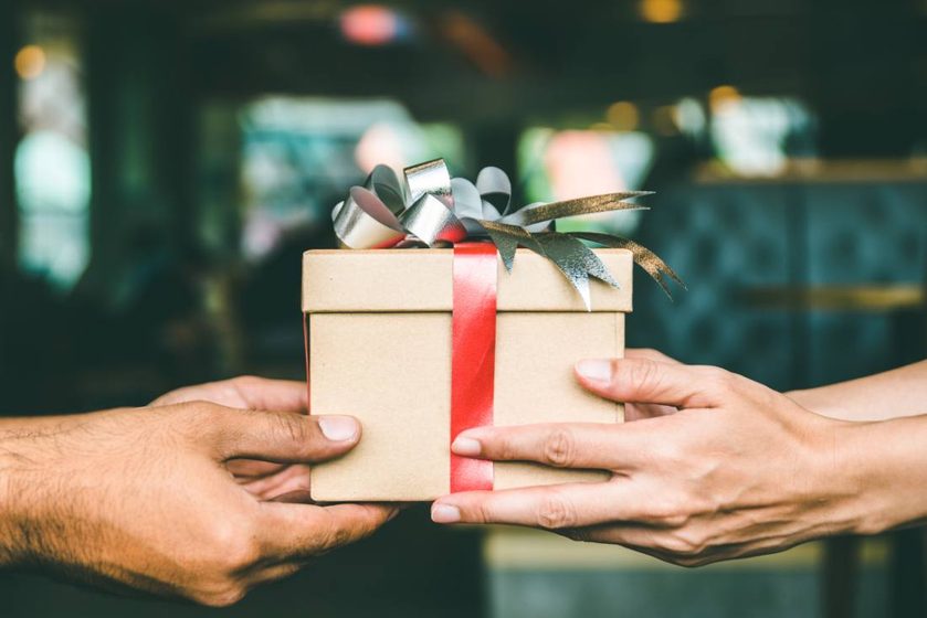 Two pairs of hands passing a gift box tied with a red ribbon.