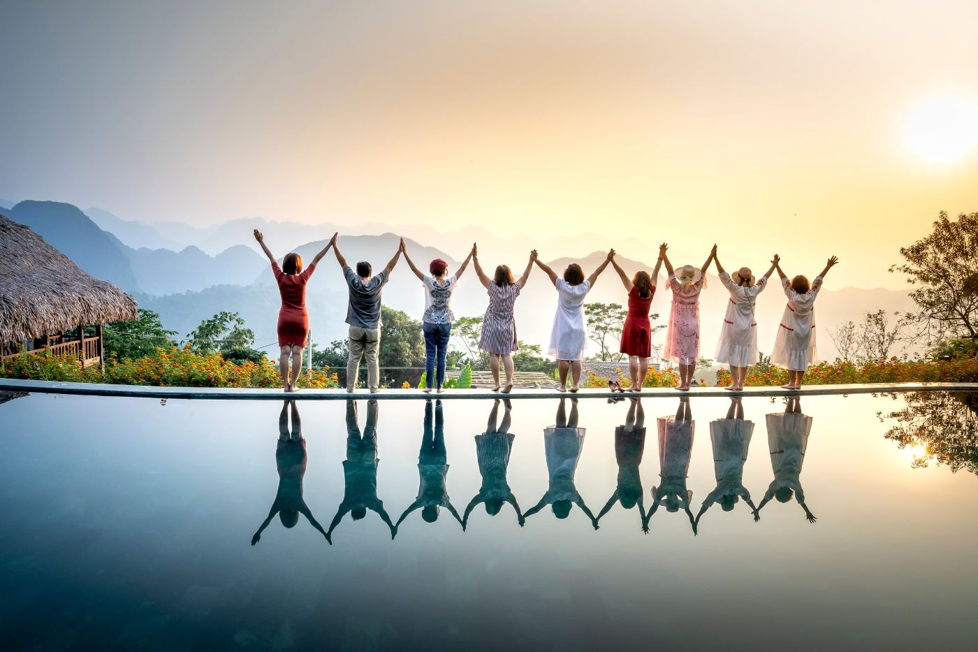 A row of people at the edge of a pool at sunset, arms raised, their reflections in the water and mountains behind.
