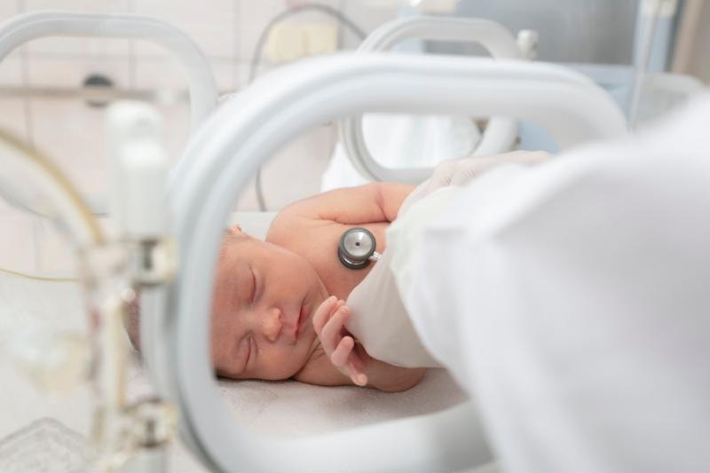 A newborn asleep in an incubator while a doctor listens to its heart.